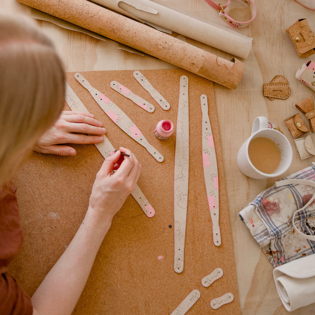Hand-painted leather collar and dog leash handles with pink details on cream beige cork, displayed on a studio table with a cup of coffee, rolls of cork leather, and other creations in the making