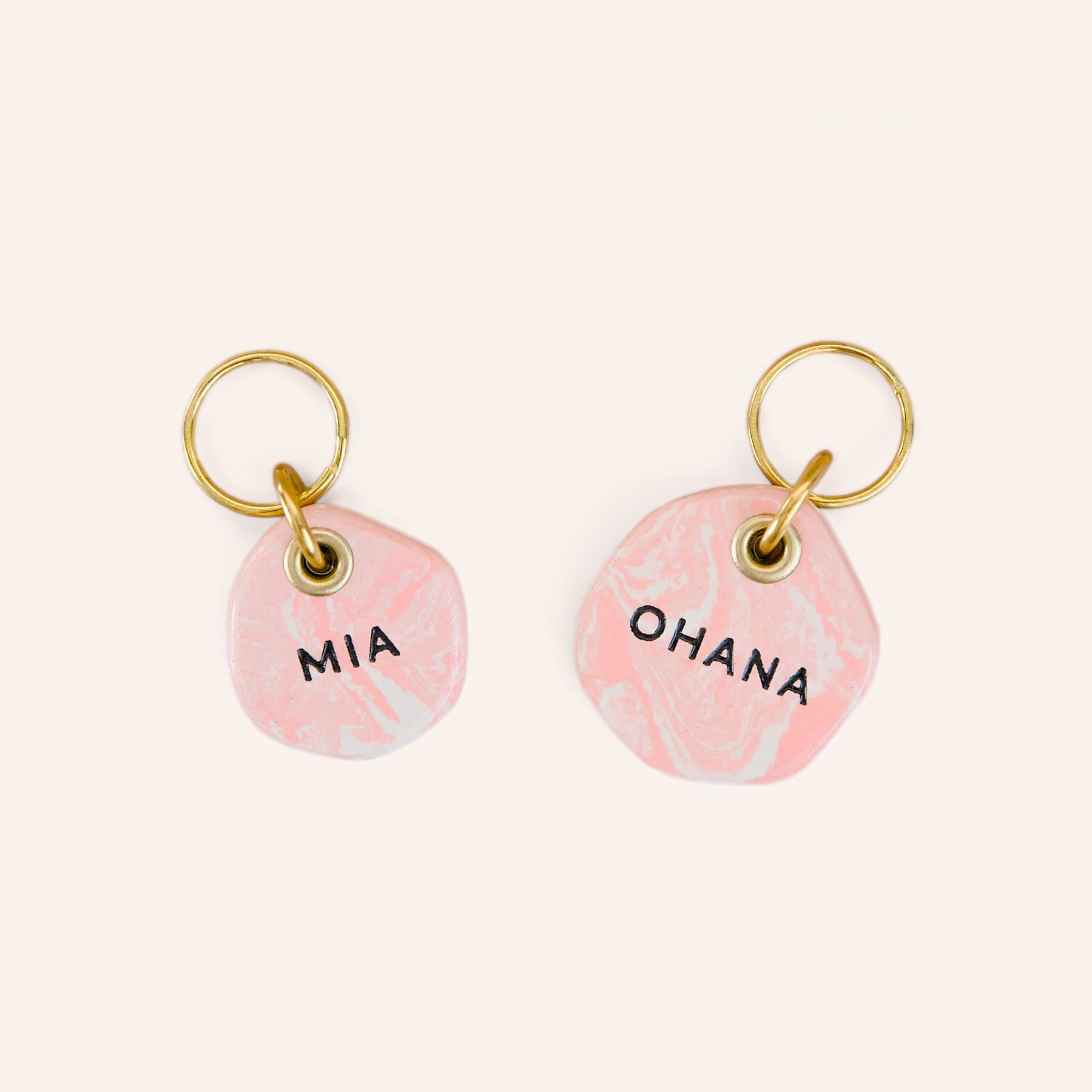 Two personalised pink marble-look clay dog name tags engraved with “Mia” and “Ohana”, with brass split rings