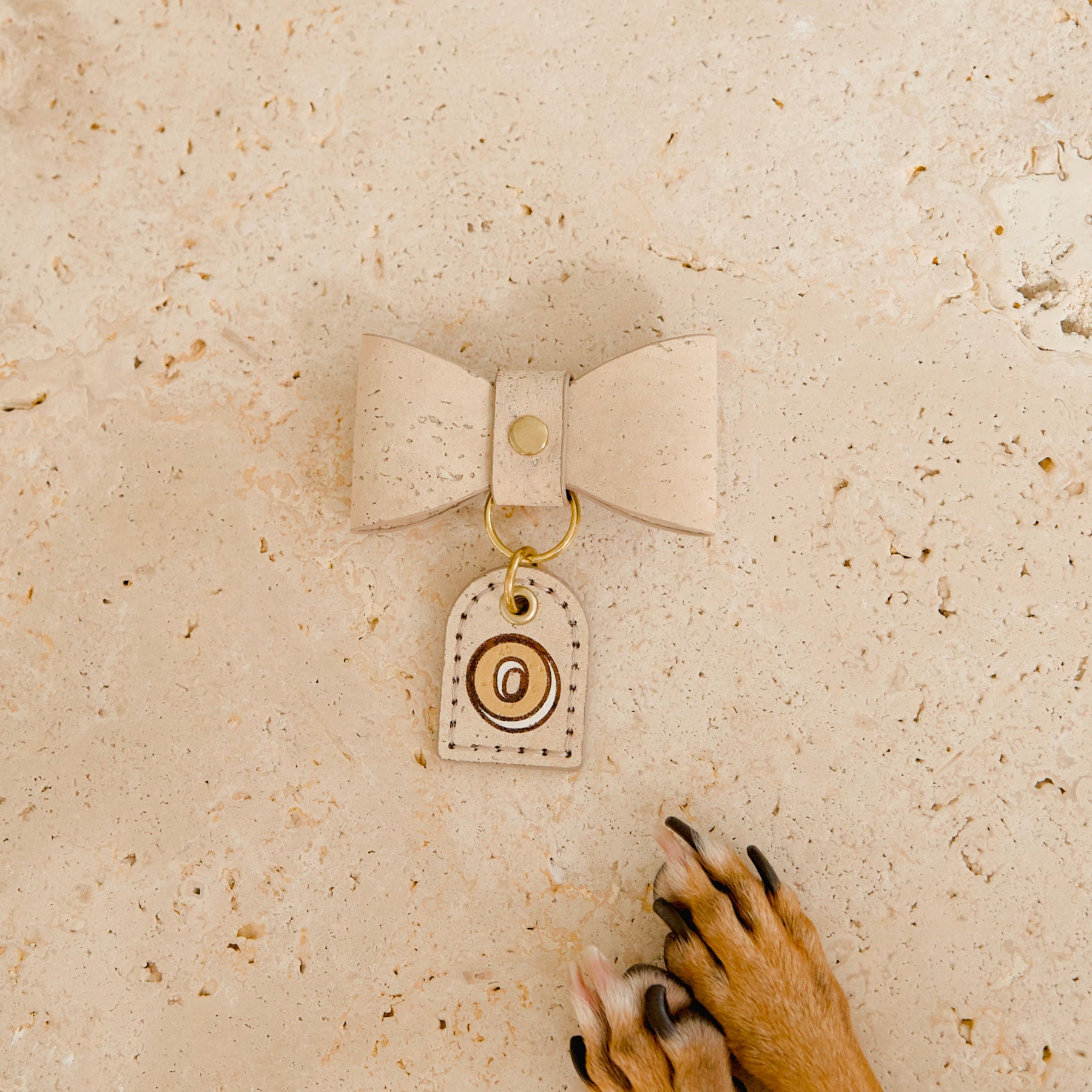 Cream cork leather dog bow tie with personalised monogram cork tag, dog paws visible, on travertine tile