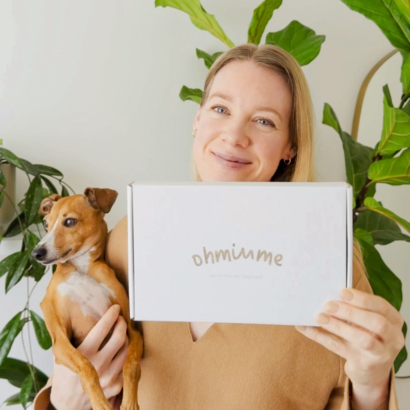 Woman holding a small dog and a box with “ohminme” branding, smiling at the camera