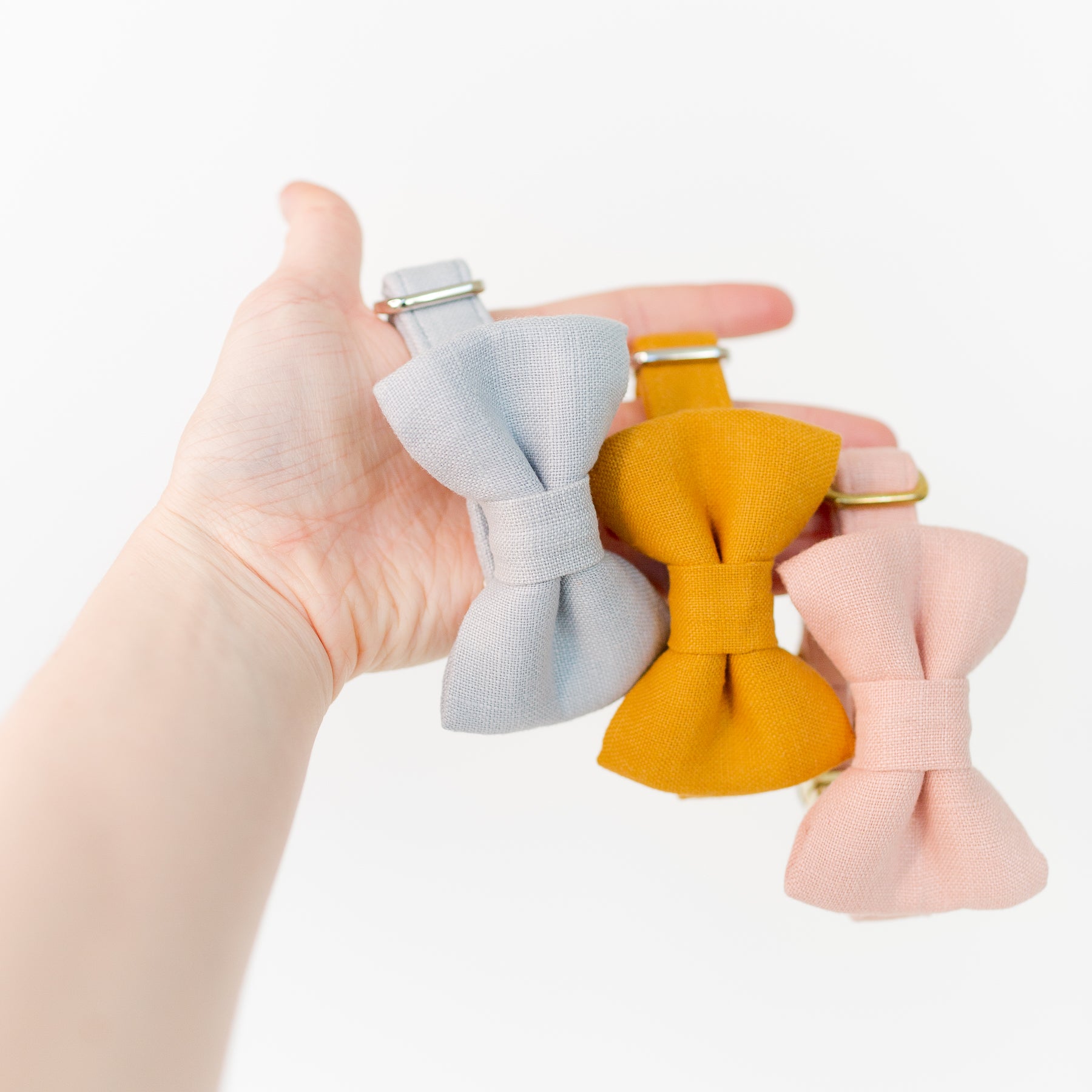 Hand holding three linen dog bow tie collars in pink, sky blue, and mustard yellow.
