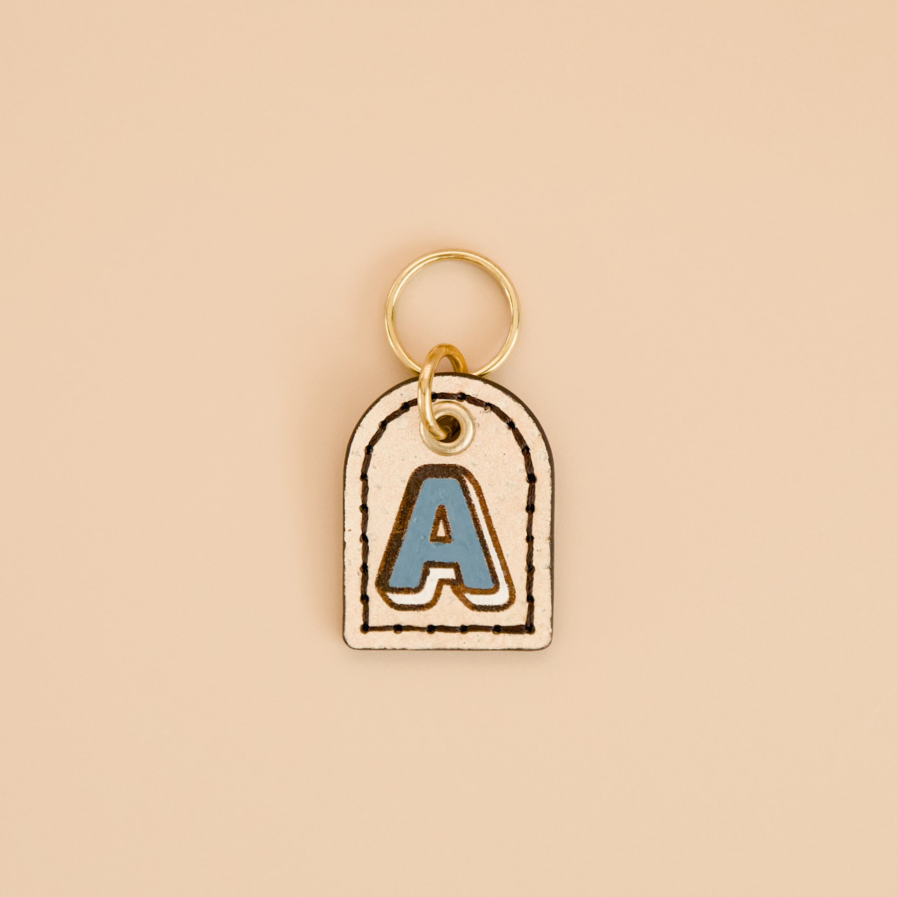 Beige cream cork leather dog ID tag with blue hand-painted monogram “A” and dark espresso brown edges